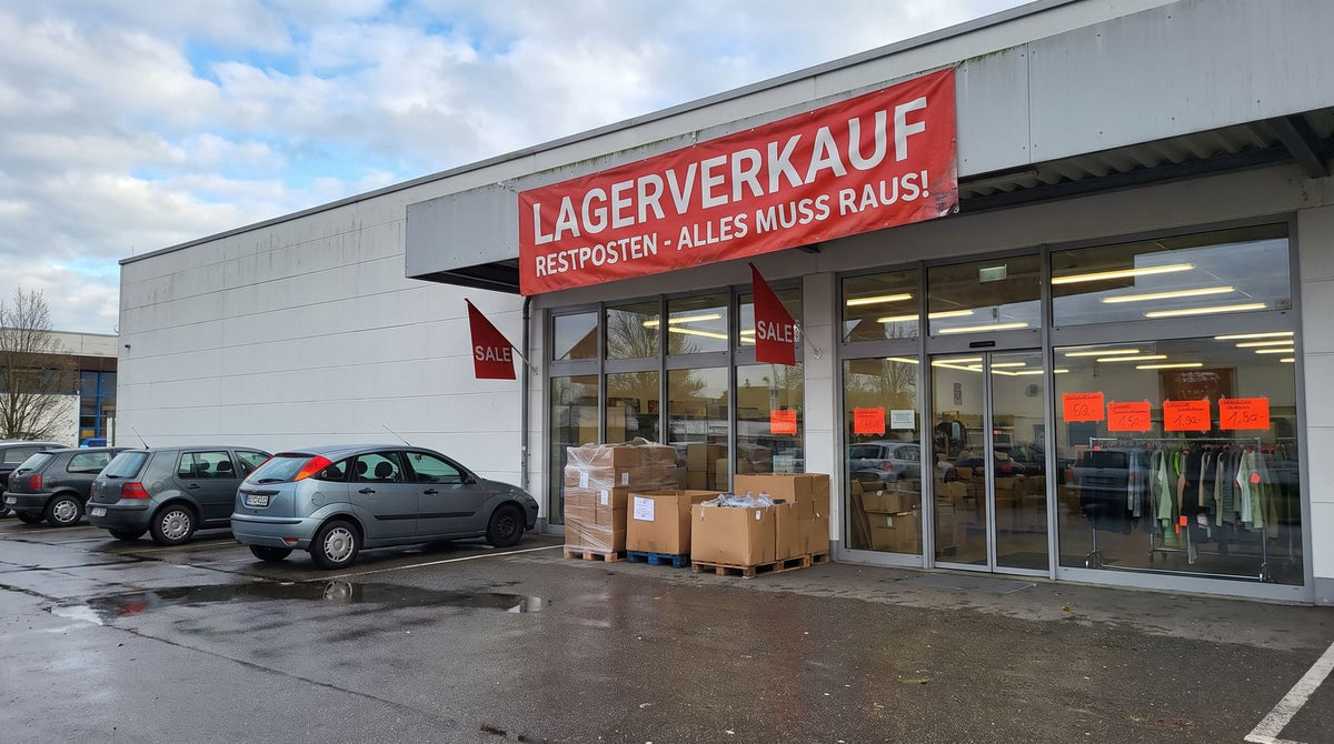 Außenansicht Lagerverkauf Manching, Weberstraße 10, mit rotem Banner „Lagerverkauf – Restposten“ und kostenlosen Parkplätzen